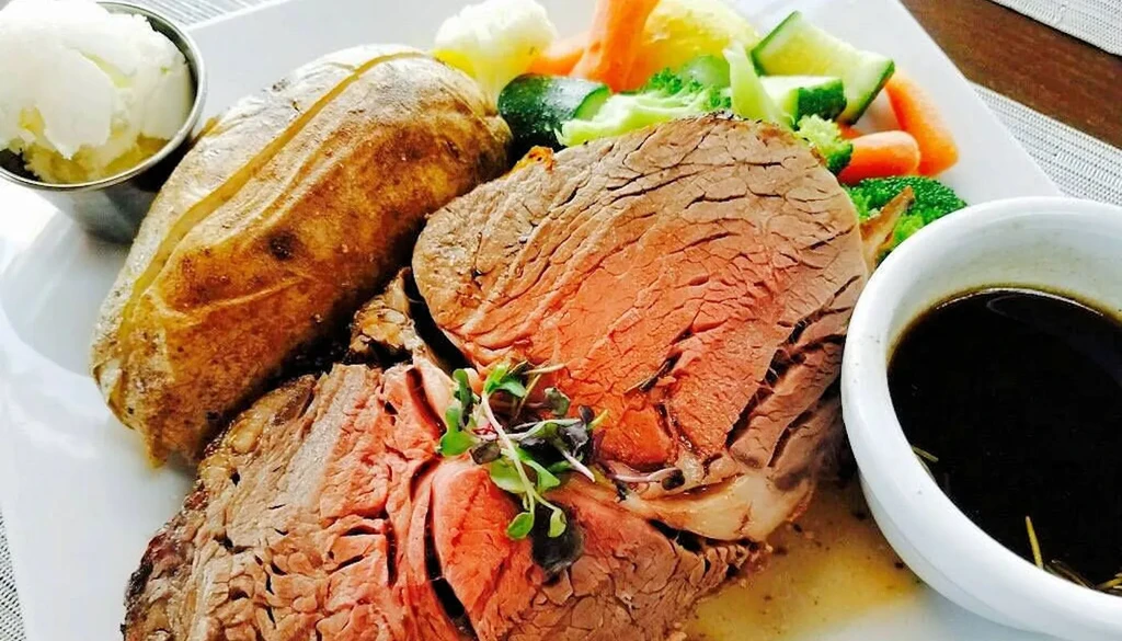 Generous serving of medium-rare roast beef, baked potato, colorful vegetables, sauce.