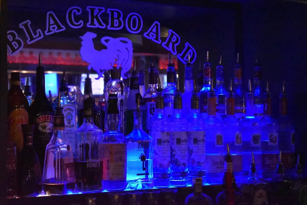 Blue-lit bar bottles with pour spouts, reflecting "BLACKBOARD" sign.