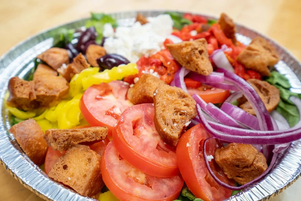 Generous salad with tomatoes, feta, olives, peppers in a foil container.