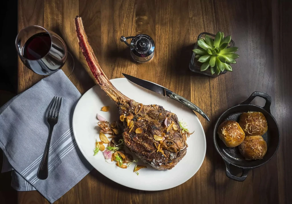 Overhead: Tomahawk steak, red wine, bread rolls on a dark table.
