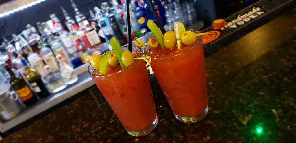 Two garnished Bloody Marys on a bar counter, liquor bottles blurred.