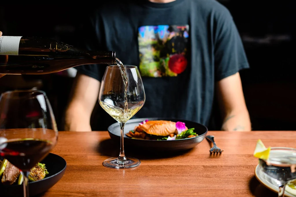 White wine pours into a glass beside salmon with a pink flower.
