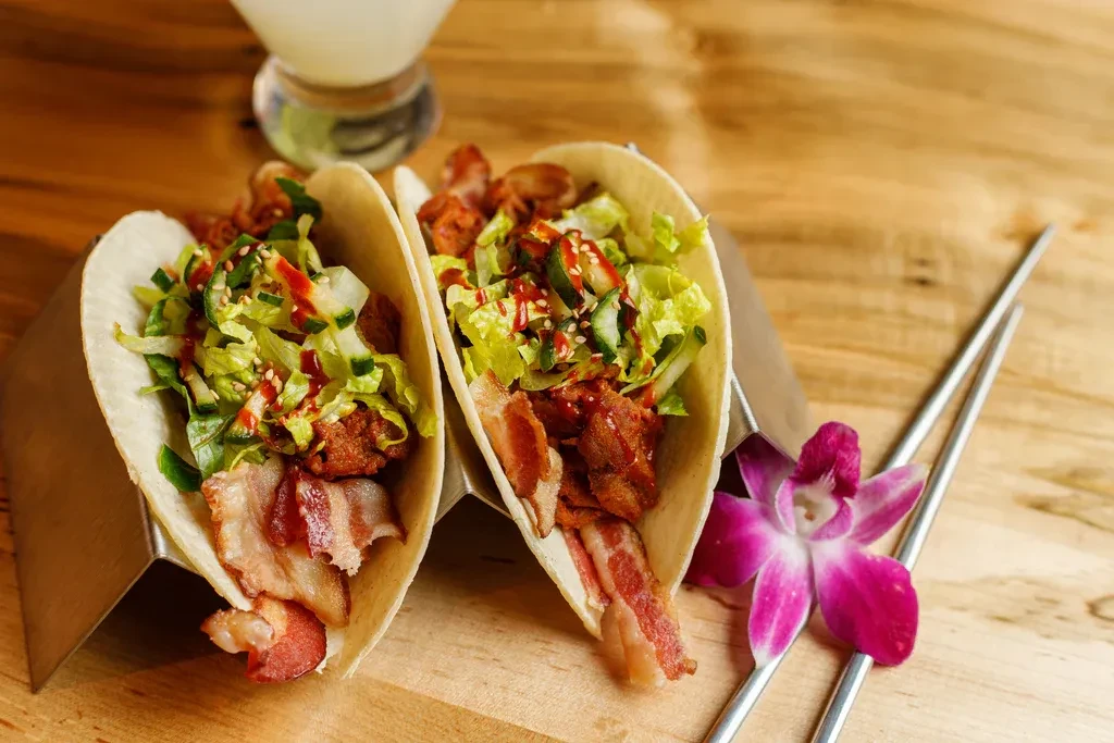 Two tacos filled with chicken, bacon, sauce, and a purple orchid.