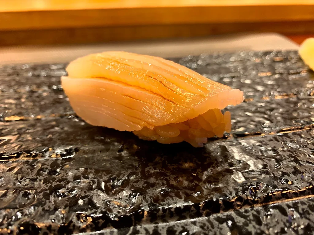 Close-up of nigiri sushi with pale shellfish topping on a dark plate.