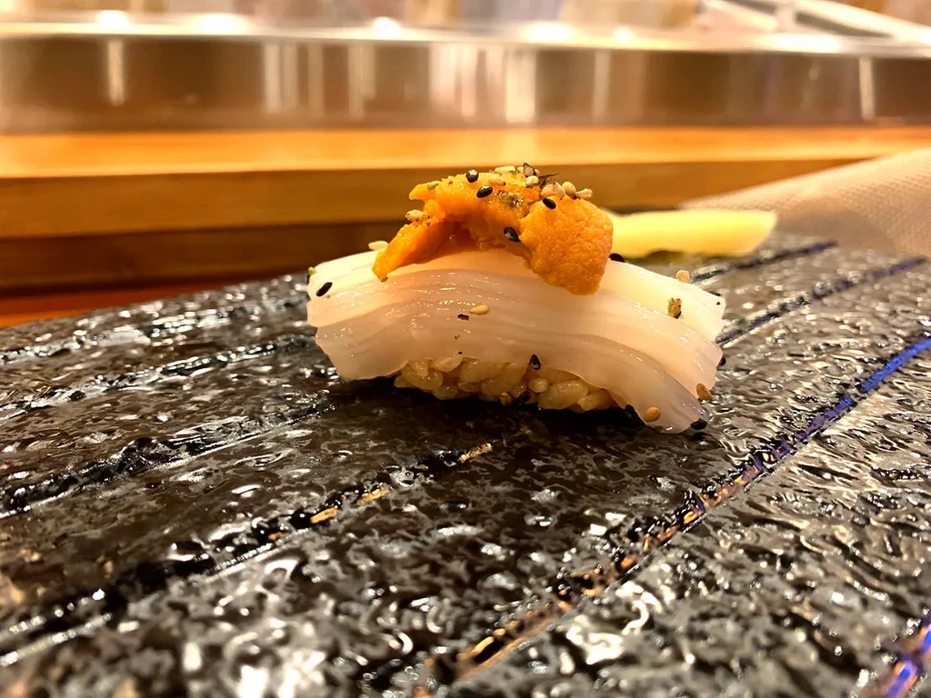 Nigiri sushi with squid, uni, and sesame seeds on a dark plate.