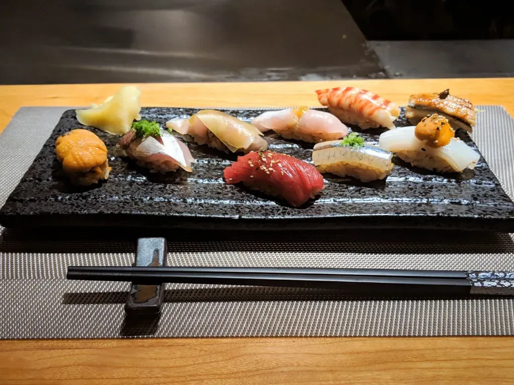 Nigiri sushi assortment on black plate with pickled ginger and chopsticks.