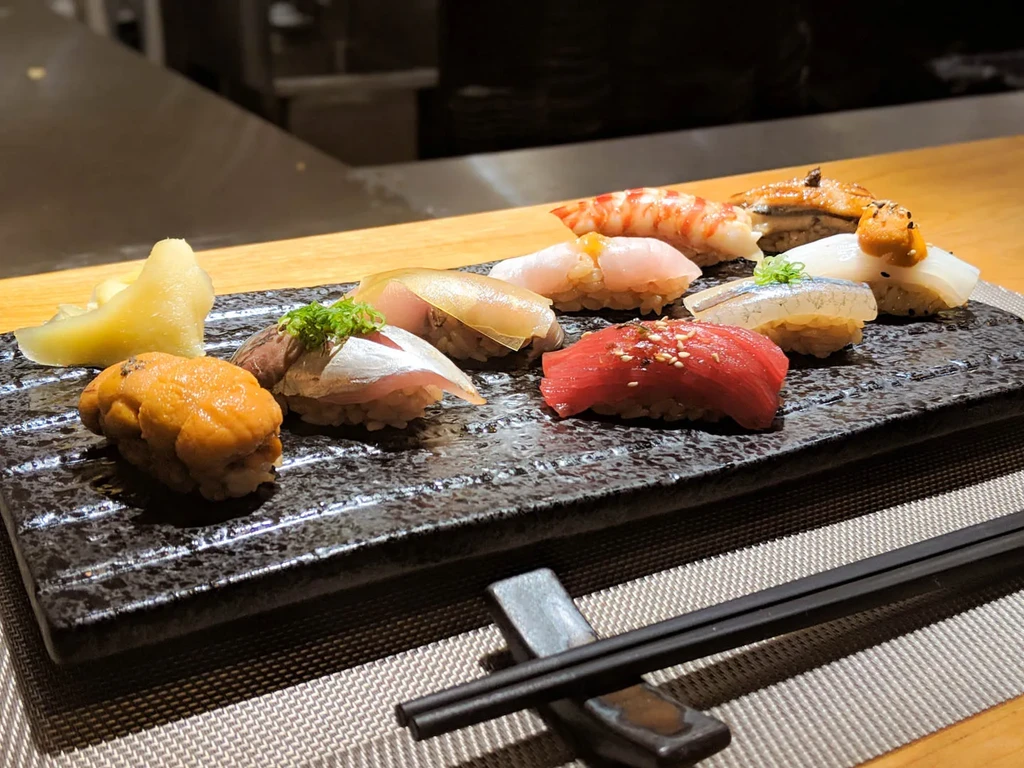 Ten diverse nigiri sushi pieces on a plate with ginger and chopsticks.