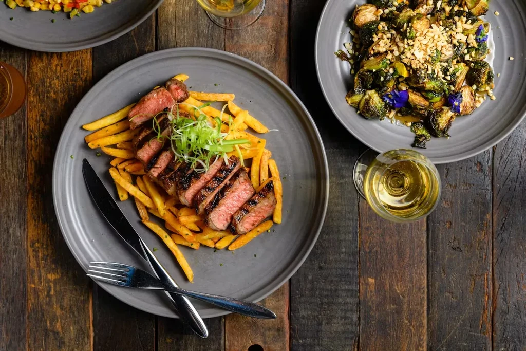 Overhead view of sliced steak, fries, brussels sprouts, wine on rustic table.