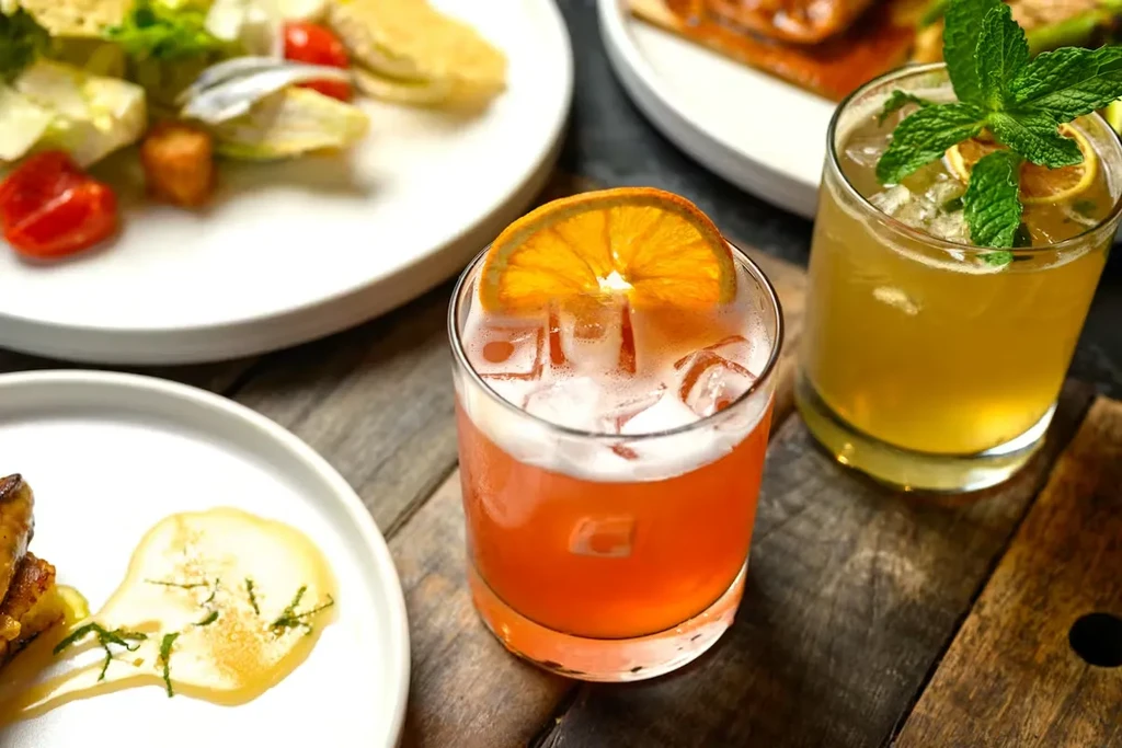 Two vibrant cocktails, salad, and sauce on a wooden dining table.