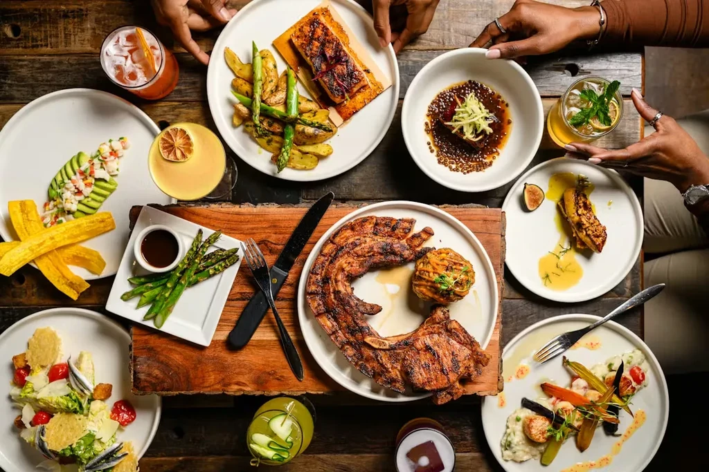 Rustic dining table flat lay with gourmet food, cocktails, and hands.