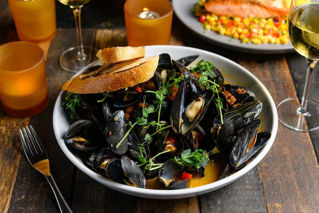 White bowl of mussels in savory broth with baguette and microgreens.