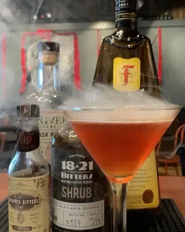 Smoky reddish-orange cocktail in martini glass, flanked by spirit and bitters bottles.