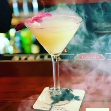 Pale yellow cocktail smoking in martini glass with a pink orchid.