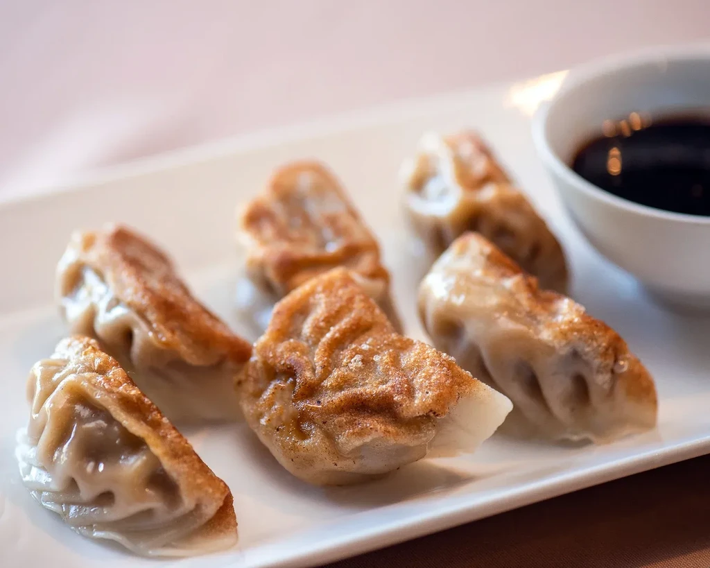Six golden-brown potstickers with dipping sauce on a white plate.