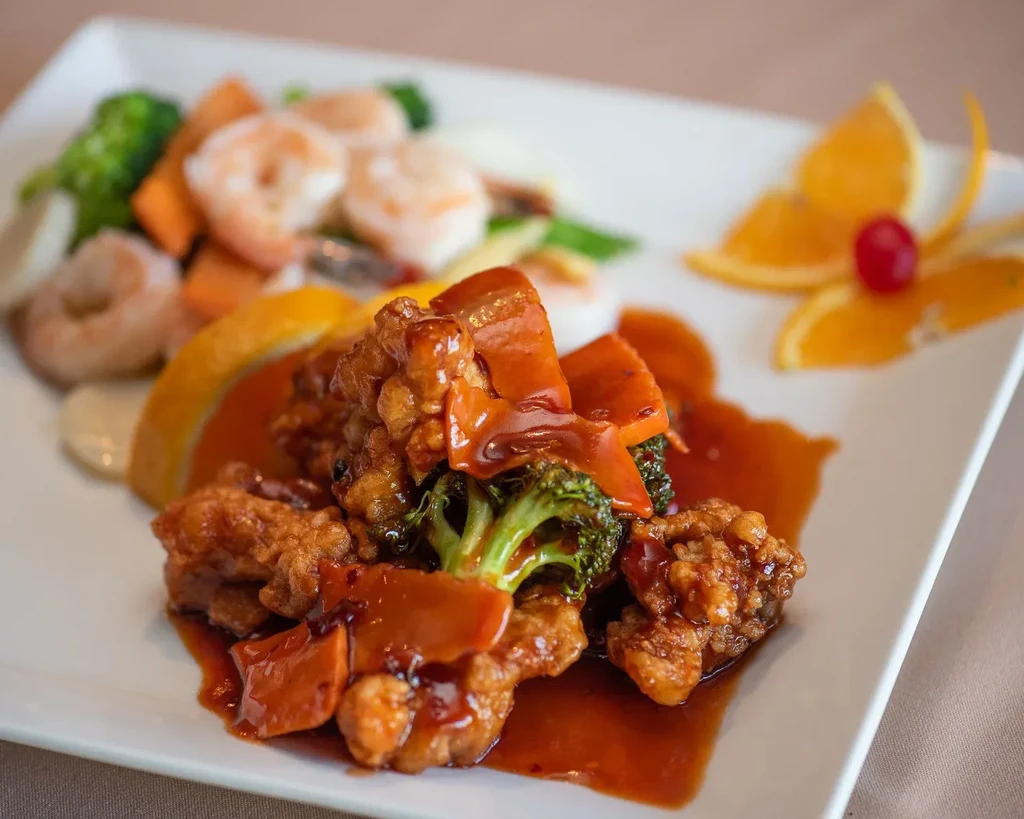 Meticulously plated orange chicken, shrimp, and vegetables with fruit garnish.