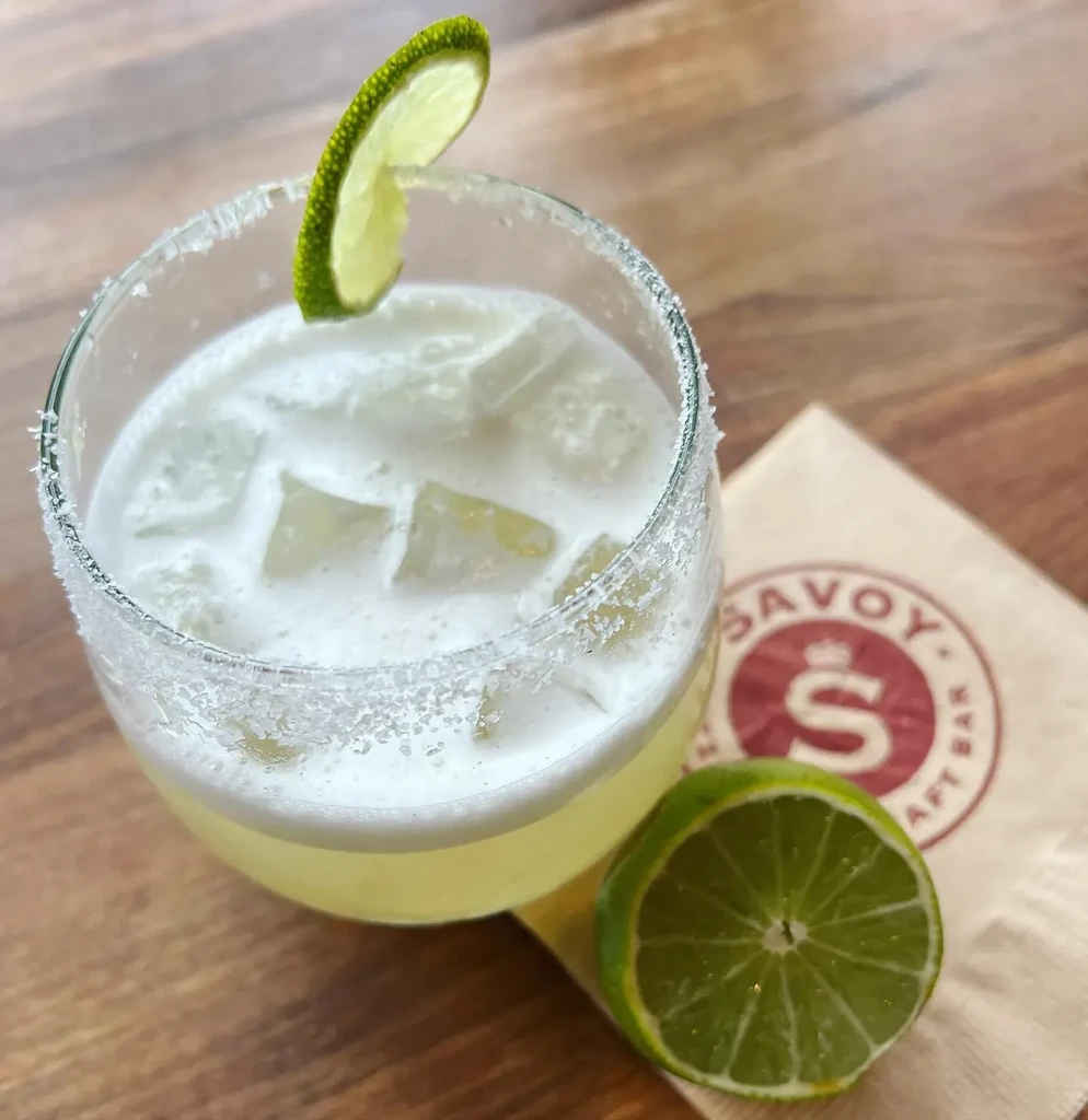 Salt-rimmed pale-green cocktail with lime slice on wooden table.