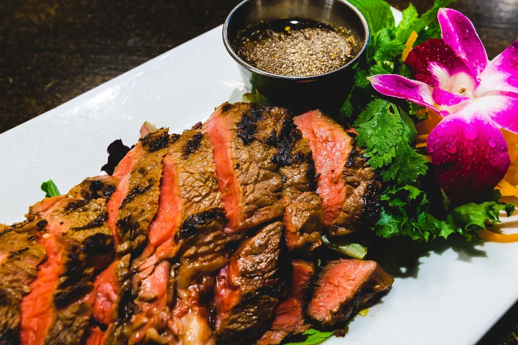 Plated grilled medium-rare steak with dipping sauce, herbs, and orchid.