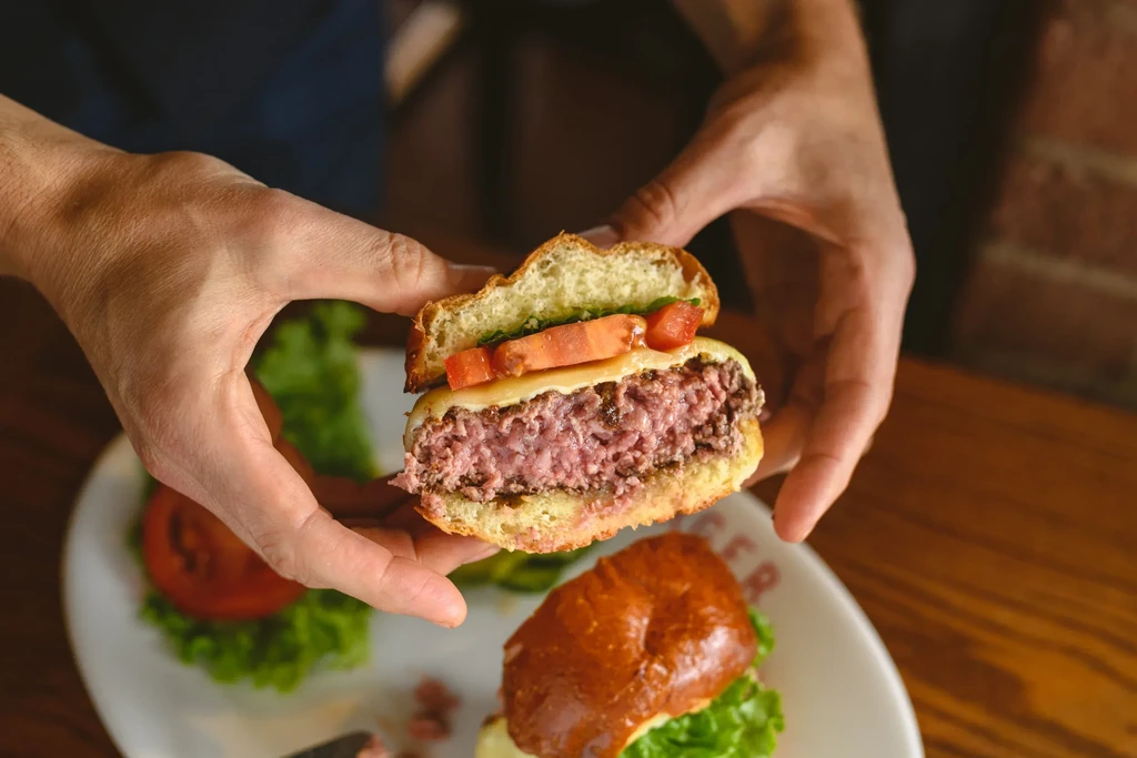 Hands hold a halved burger, revealing medium-rare patty, cheese, tomato, sauce.