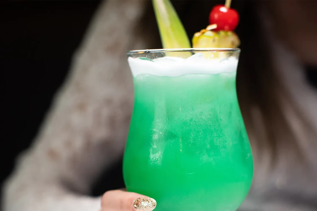 Frosty green tropical cocktail with pineapple, cherry garnish, held by a hand.