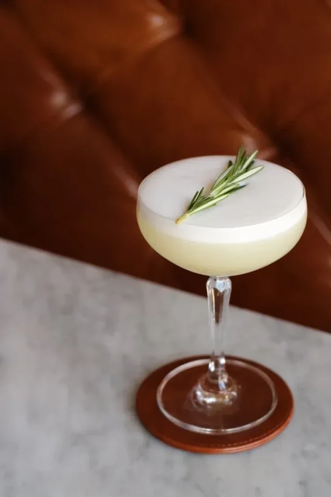 Cocktail with white foam and rosemary, on leather coaster and marble.