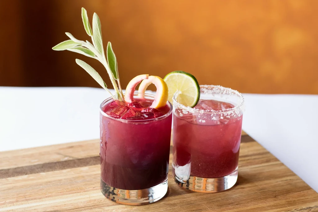 Two cold reddish-purple cocktails garnished with herbs and citrus on wood.