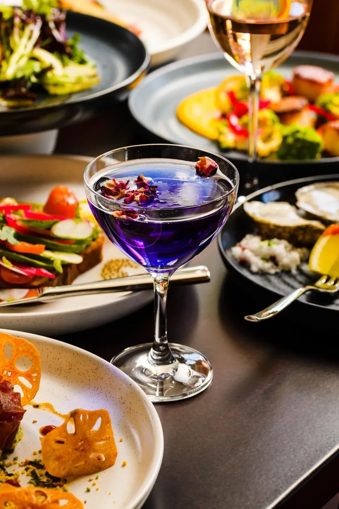 Restaurant table laden with a purple cocktail and diverse food dishes.