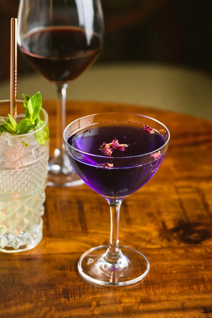 Purple cocktail, clear drink with mint, and red wine on wood table.
