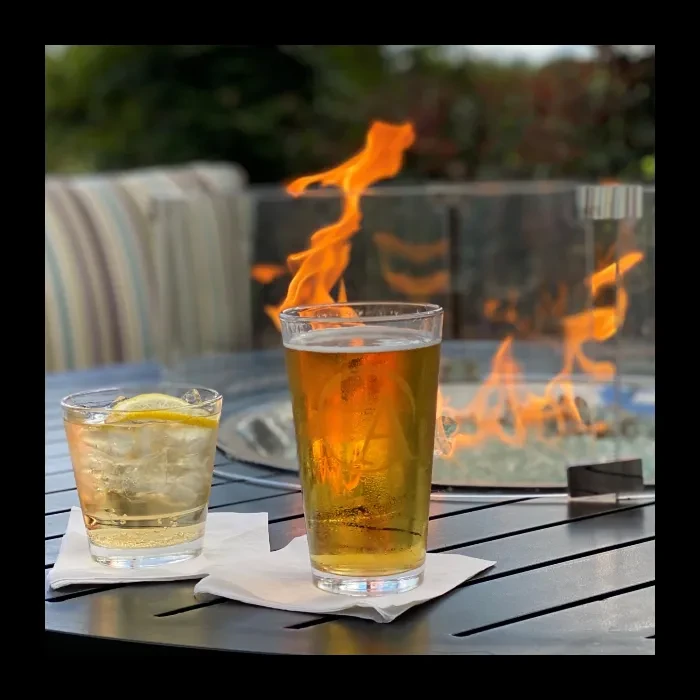 Two drinks, beer and lemon, on an outdoor table by a glowing fire pit.