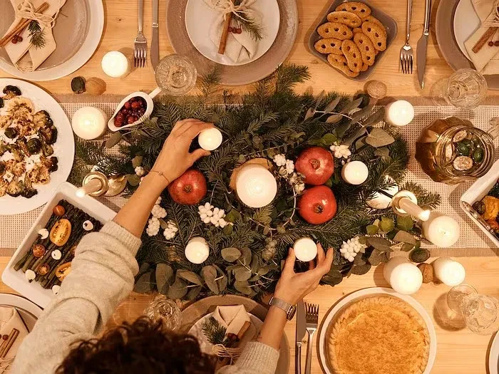 Festive dining table with garland centerpiece, food, and hands adjusting a candle.