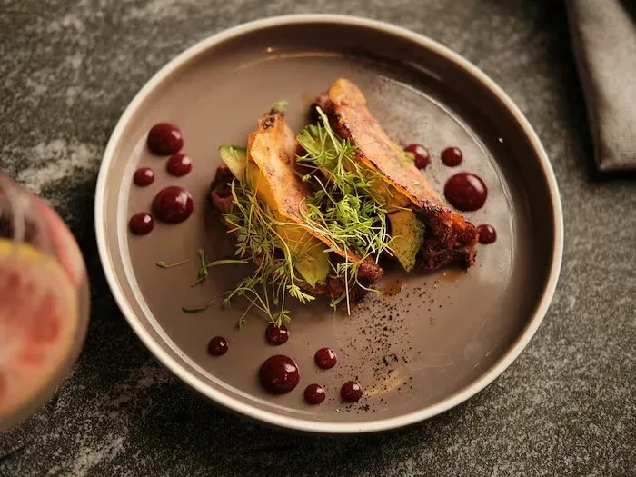 Plated gourmet dish: roasted food, microgreens, dark sauce, citrus drink.