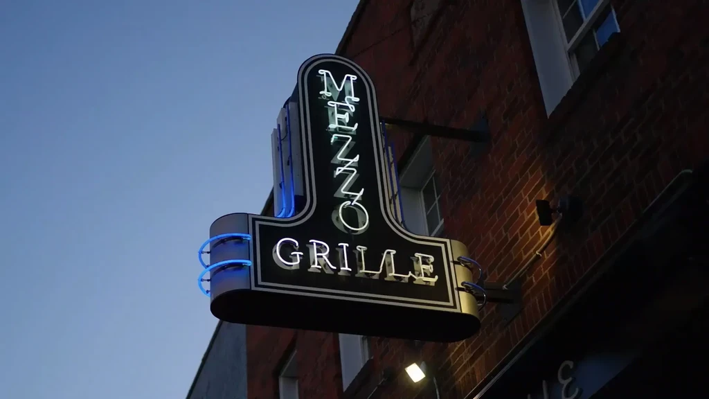 Art deco 'MEZZO GRILLE' neon sign, white and blue, on brick.