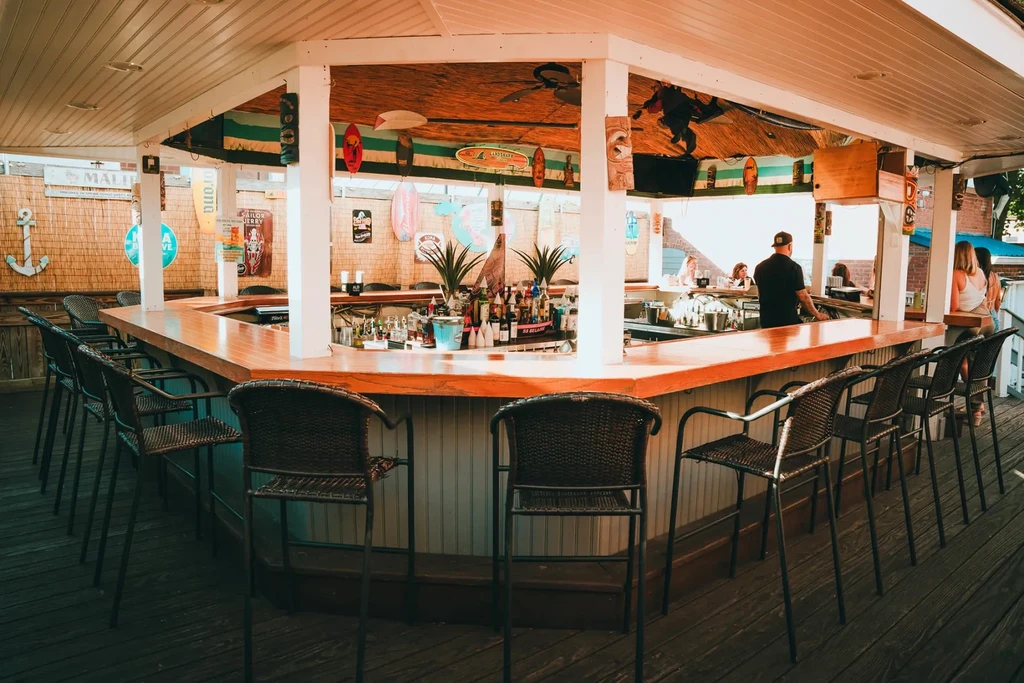 Open-air tropical bar with U-shaped counter, patrons, and surf decor.