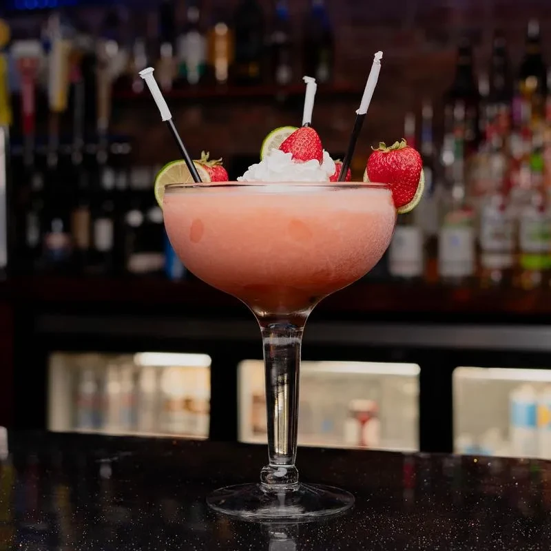 Pink frozen cocktail with cream, strawberries, lime, and straws on bar.