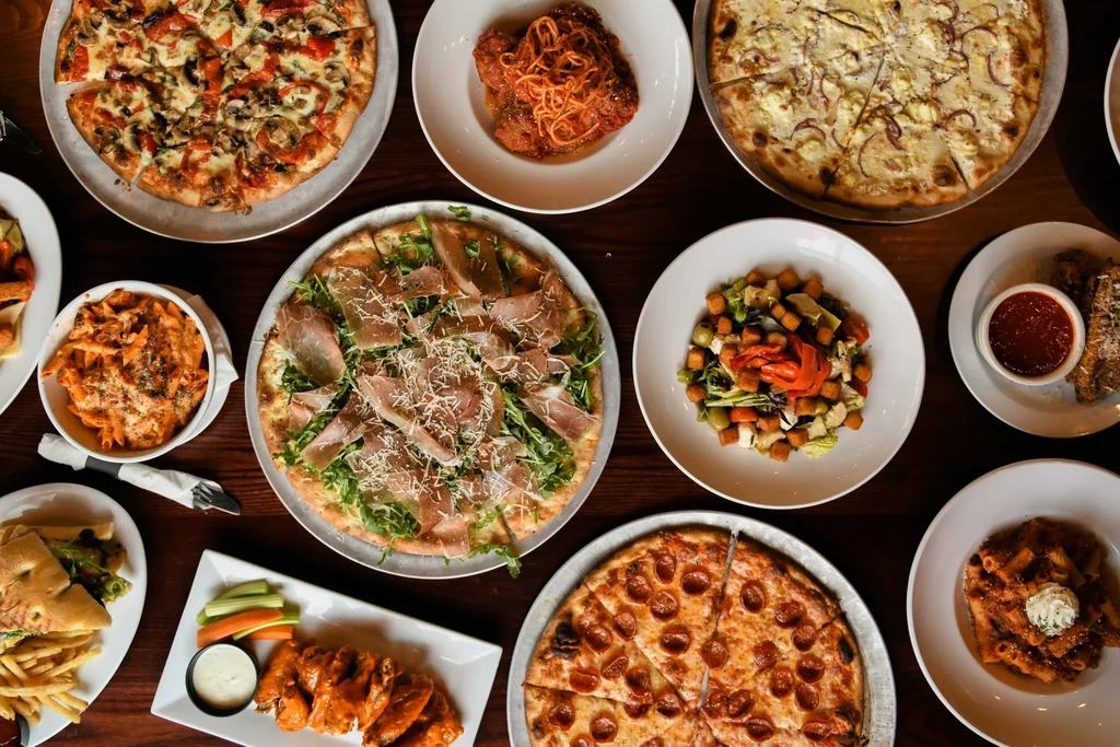 Overhead view: wooden table piled with diverse Italian and American dishes.