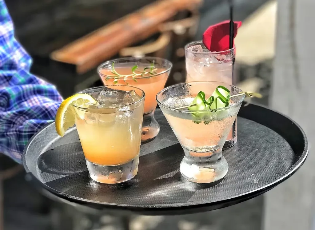 A person holds a tray of four colorful, garnished cocktails.