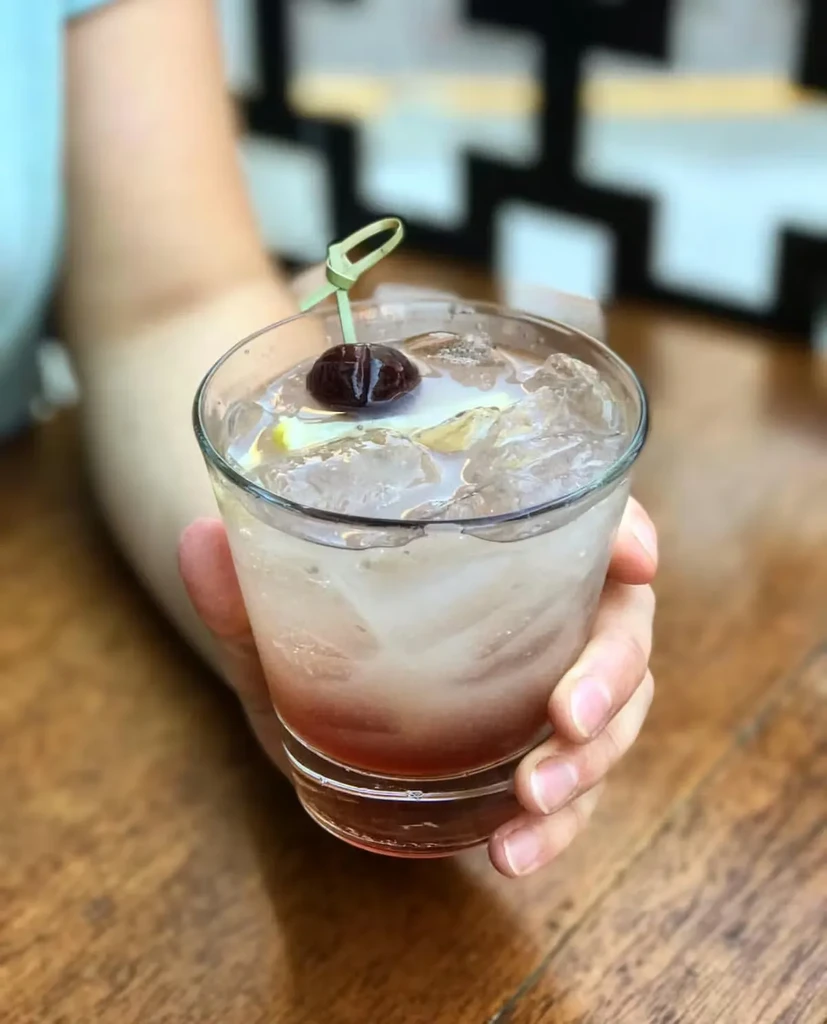 Hand holding a gradient red cocktail with cherry and lemon.