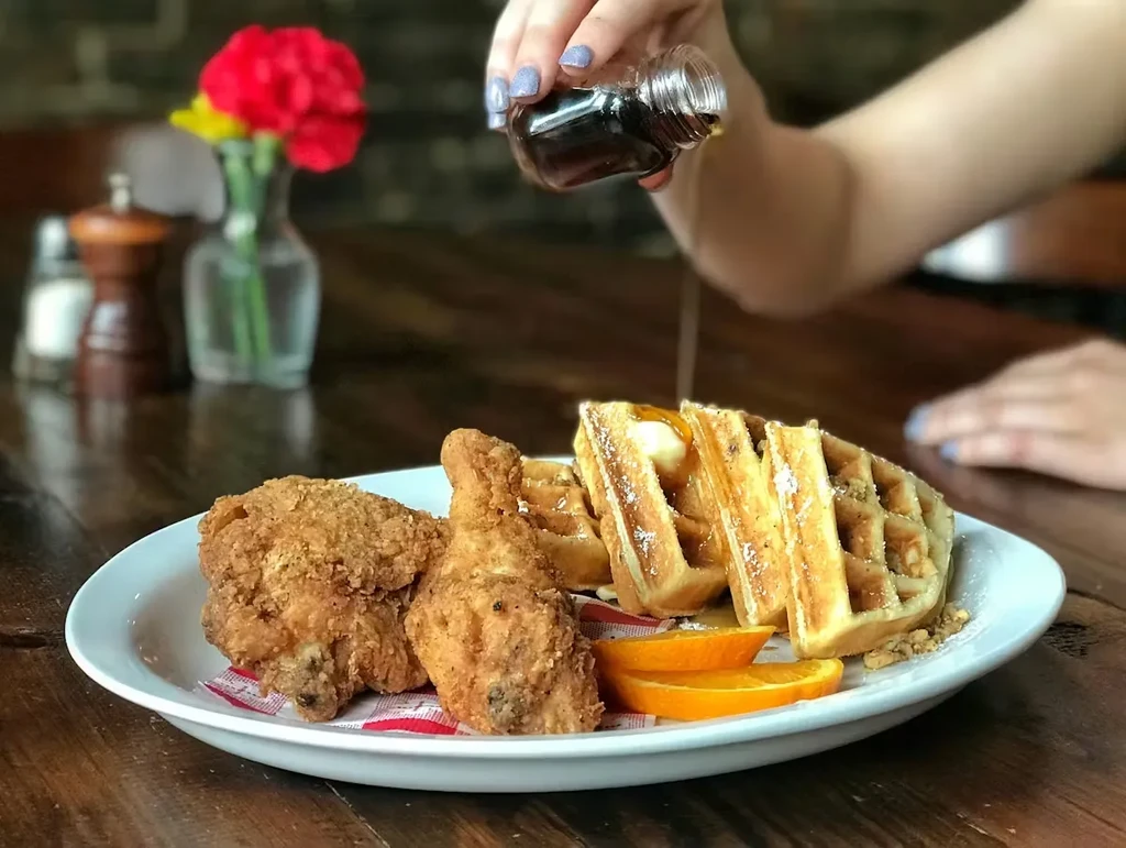 Hand pouring syrup on chicken and waffles on a rustic wooden table.