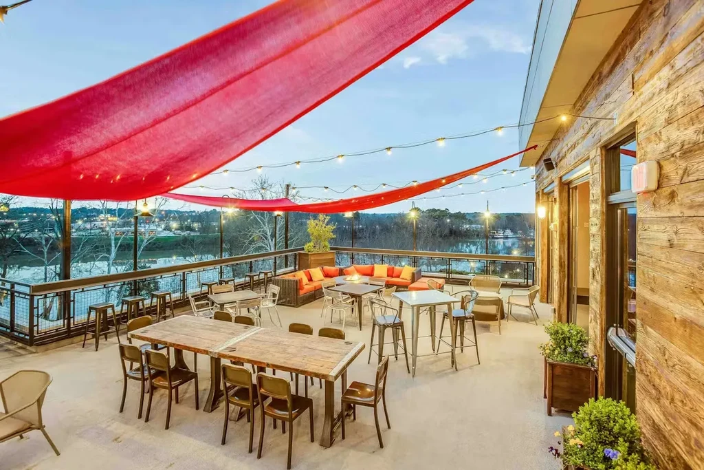 Outdoor restaurant patio with red shades, string lights, diverse seating, overlooking water.