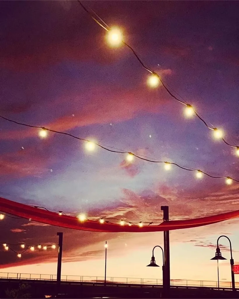 Vibrant sunset sky illuminates glowing string lights, a red banner, and pier.