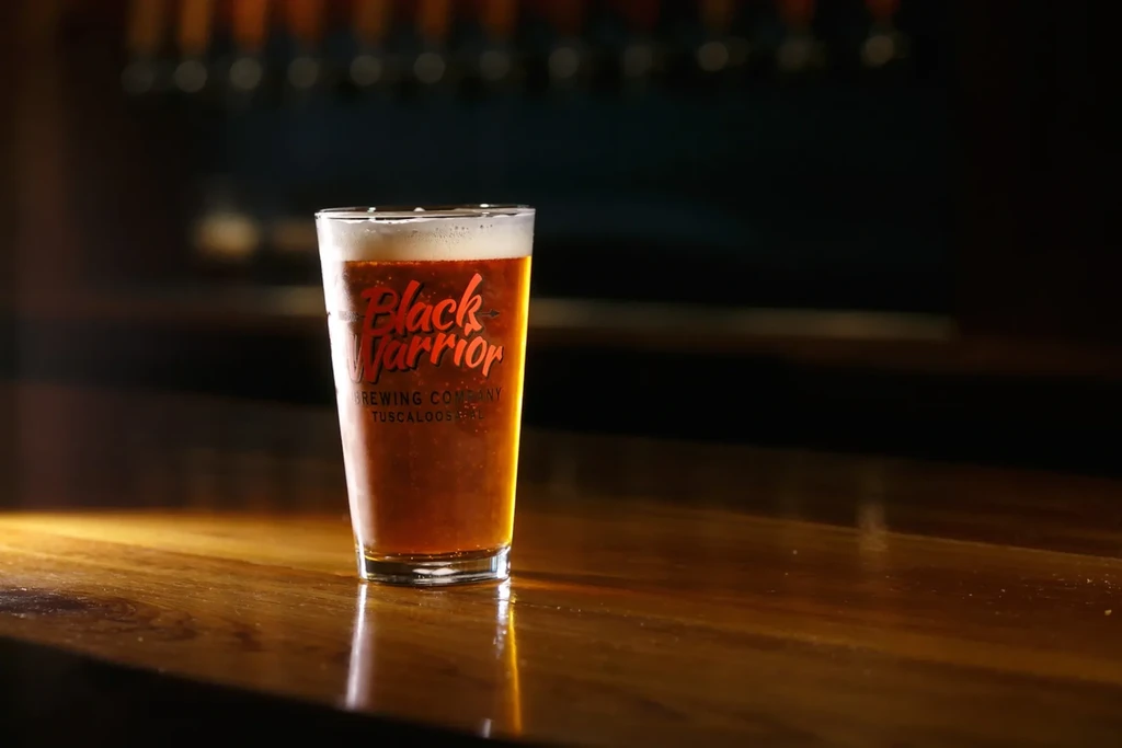 Pint glass of amber beer branded Black Warrior Brewing Company on a wooden bar.