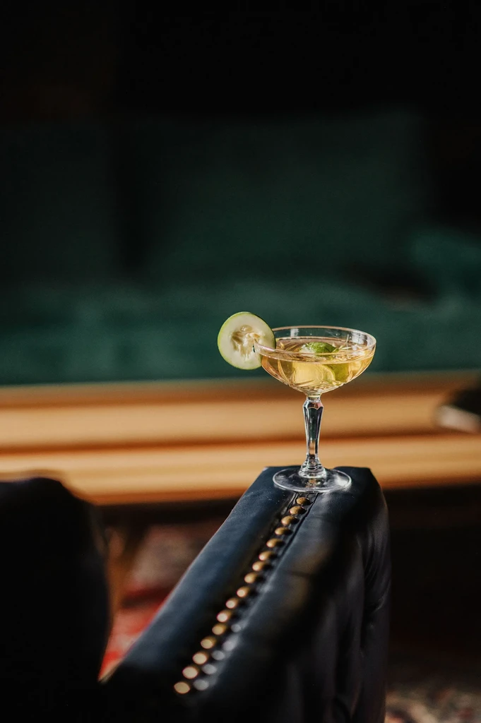 Clear yellowish cocktail in coupe with cucumber, mint on studded furniture.