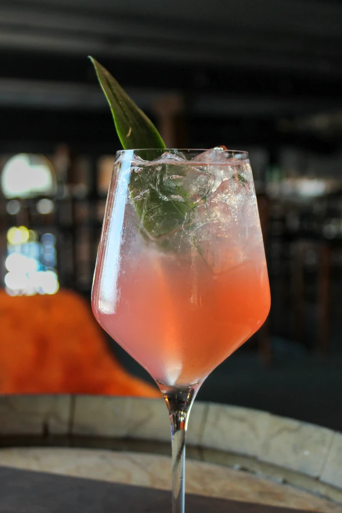 Pink-orange cocktail with ice and a green leaf in a stemmed glass.