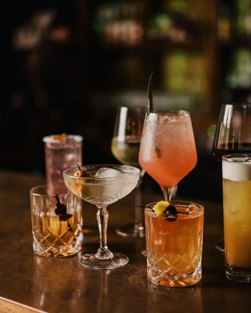 Seven colorful cocktails in diverse glasses on a dark bar counter.