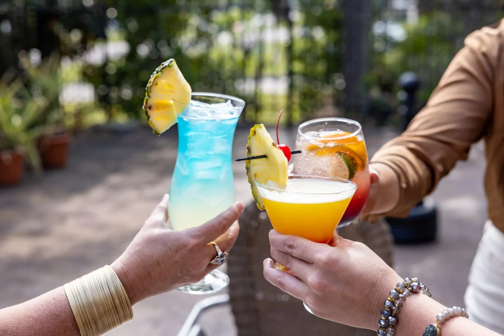 Three hands raise colorful, fruit-garnished cocktails outdoors.