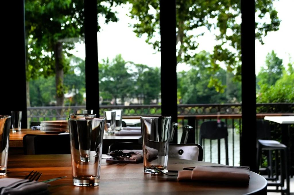 Empty restaurant table with glasses by windows overlooking water and trees.