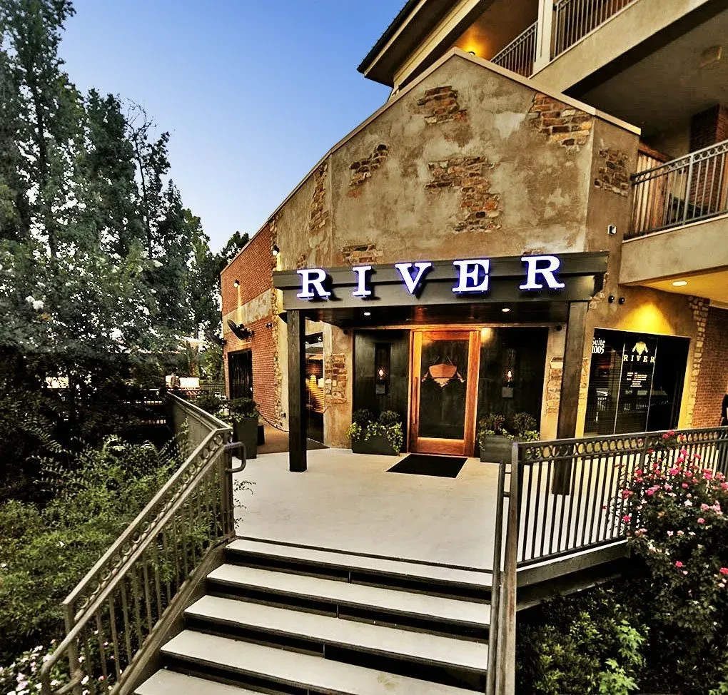 Illuminated "RIVER" sign, brick building, concrete stairs, and plants.