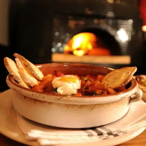 Rustic stew with poached egg, baguette, and glowing wood-fired oven.
