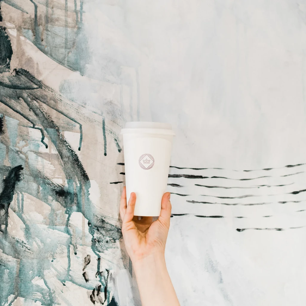 Hand holds white coffee cup with crown logo against abstract mural.