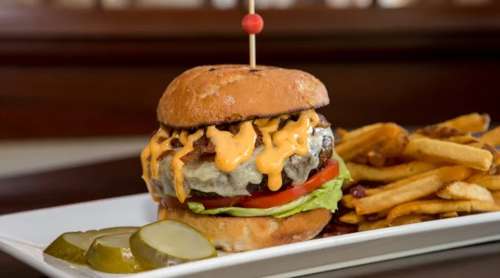 Stacked gourmet burger with cheese, bacon, fries, pickles, and skewer.