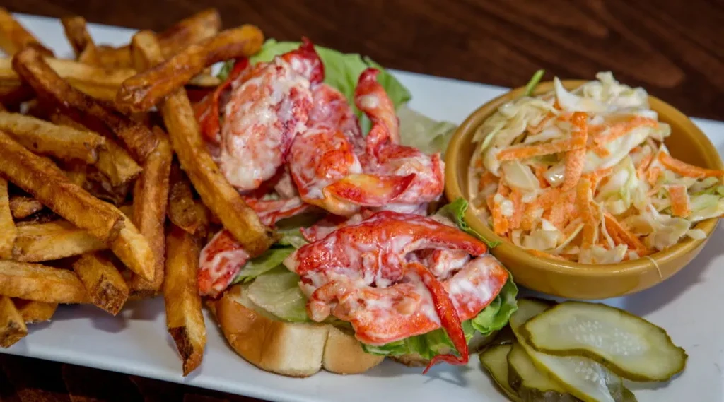Lobster roll with french fries, coleslaw, and pickles on a white plate.